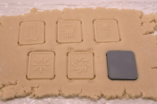 Five cookie designs are shown pressed into rolled out dough, and a plastic mold is pressed into the dough for a sixth cookie