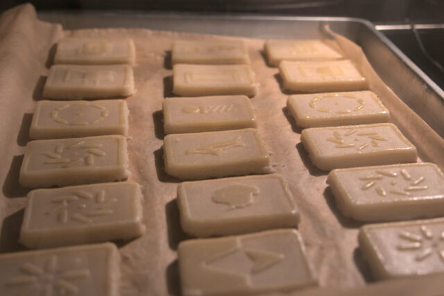 Twelve cookies with raised designs are on a cookie sheet baking in an oven