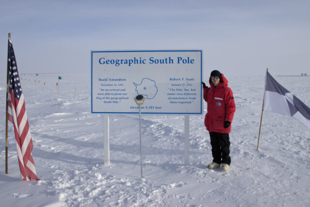 Large white "Geographic South Pole" sign installed in snow with person wearing red parka standing next to it and metal Geographic South Pole marker in front of it.