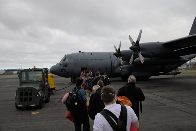 A line of people waiting to board a C-130 on the tarmac through its front door.