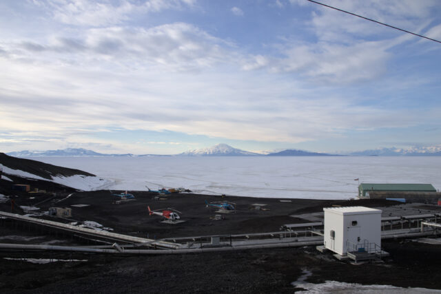 A partly cloudy sky and snow-capped mountains are visible across an expanse of sea ice, with a dirt heliport with multiple helicopters in the foreground.
