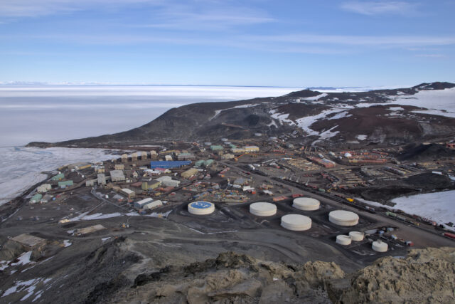 The buildings of a small town, McMurdo Station, dot a dirt-covered landscape, with sea ice to the left and some snow cover to the right. The top of a large fuel tank in the foreground is painted with the NSF logo.