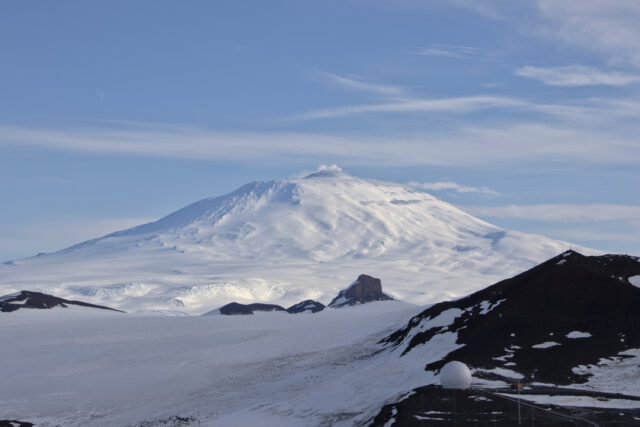 A snow-capped volcano with steam rising from the top is visible in the distance. Snow-free areas of rock and dirt are closer, with a round white radome.