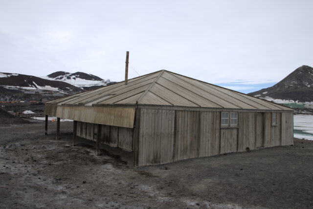 A square wooden hut with a sloped roof, surrounded by dirt, with some snow and ice in the background.