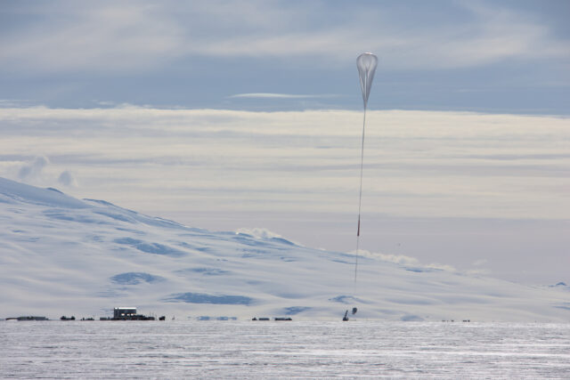 A long flight train consisting of a large high-altitude balloon and a parachute extends down to the SPIDER payload, which has just been released by the nearby launch vehicle. People, helium tanks, and the Long Duration Ballooning facilities are visible in the distance, with a flat ice shelf in the foreground, and the snow-covered slopes of Mt. Terror in the background.