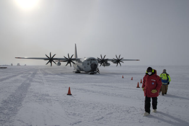 Two people walk toward the camera and away from a C-130 aircraft, parked on flat compacted snow, with its props spinning.
