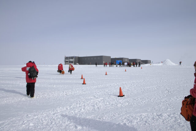 People in red parkas follow a line of orange cones along the snow toward a group of people waiting in front of the blue-gray elevated South Pole Station.