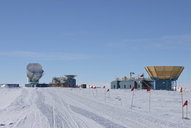 A line of flags extend along a snow road toward blue-gray buildings with exterior stairs and railings. The closer building toward the right, MAPO, has a large plywood bowl on its roof, and the building toward the left, DSL, has a metal bowl on the roof toward the right and a large telescope primary reflector to the left.
