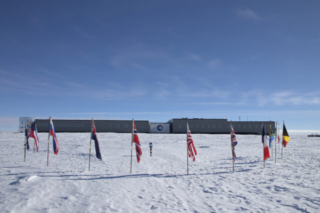 A red-and-white striped pole with a mirror ball on top is in the snow and surrounded by a set of country flags. The elevated South Pole Station is in the background.