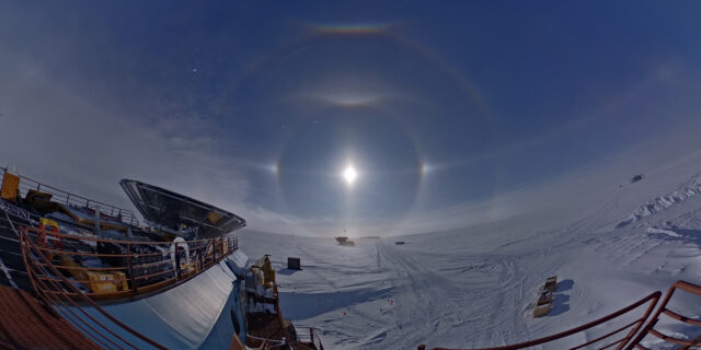 The sun is visible over a snow-covered landscape, with several rainbow-colored rings and white bright spots surrounding it. The roof of DSL and the BICEP3 ground shield are visible in the foreground.