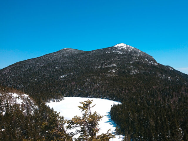 Two fir-tree-covered mountain peaks, which look like horns, are in the distance behind a snow-covered pond