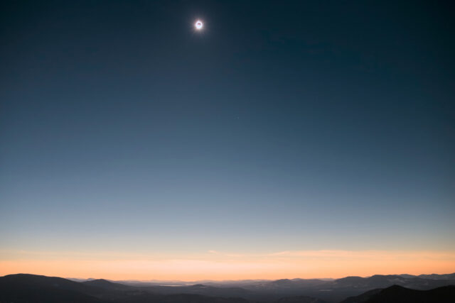 A bright light is visible peaking out of the edge of the doughnut-like moon-covered sun at the end of totality, above a brightening sunset-like sky and darkened mountains