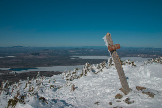 A signpost, with snow sticking to it, reads "Appalachian Trail, Biglow Mtn. - West Peak, Elev. 4150ft, M.A.T.C." Mountains and snow-covered lakes are visible in the background.