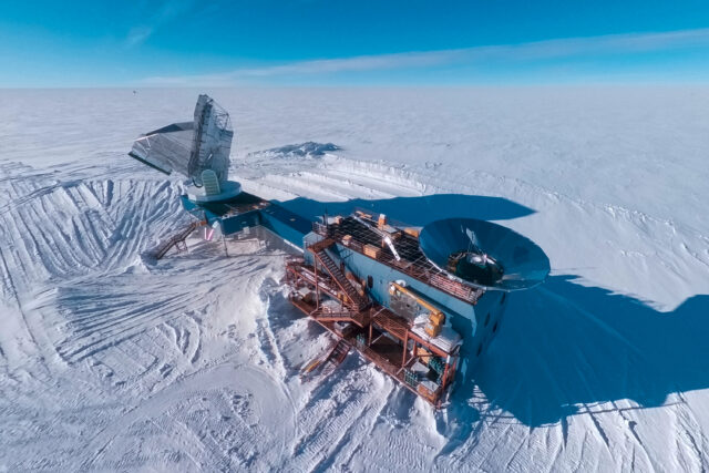 Aerial view of a two-story blue building on an ice sheet, with stairs in the front, a circular metal cone-shaped ground shield on the roof, and a large radio telescope connected to it.