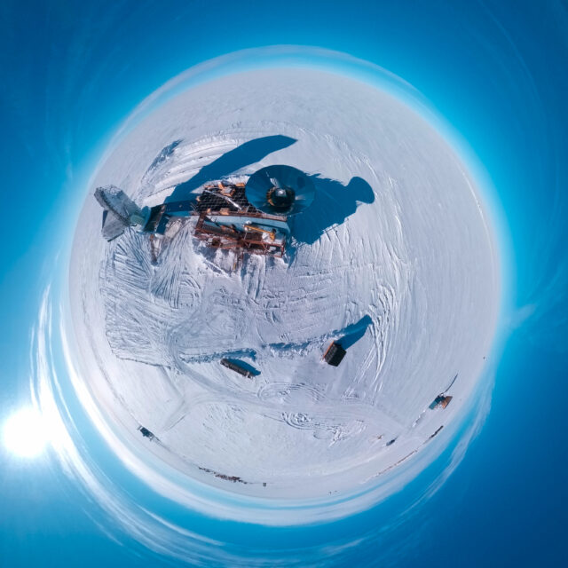 Aerial "little planet" view of a two-story blue building on an ice sheet, with stairs in the front, a circular metal cone-shaped ground shield on the roof, and a large radio telescope connected to it.