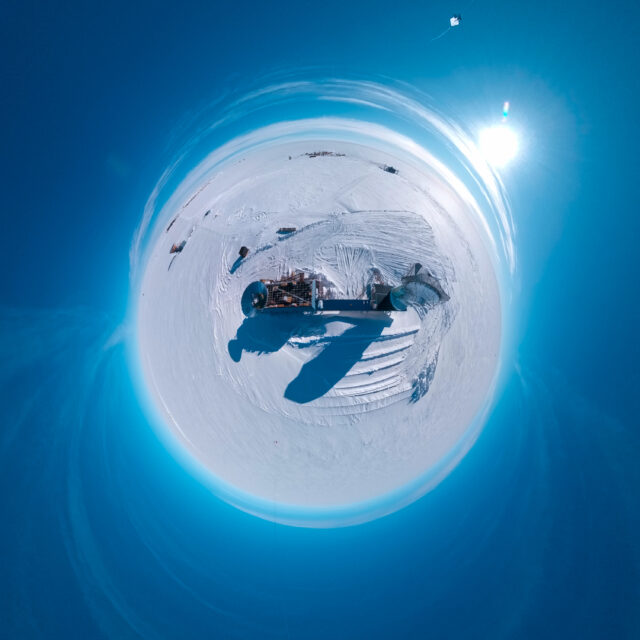 Aerial "little planet" view of a two-story blue building on an ice sheet, with stairs in the front, a circular metal cone-shaped ground shield on the roof, and a large radio telescope connected to it. A kite is visible in the corner of the image.