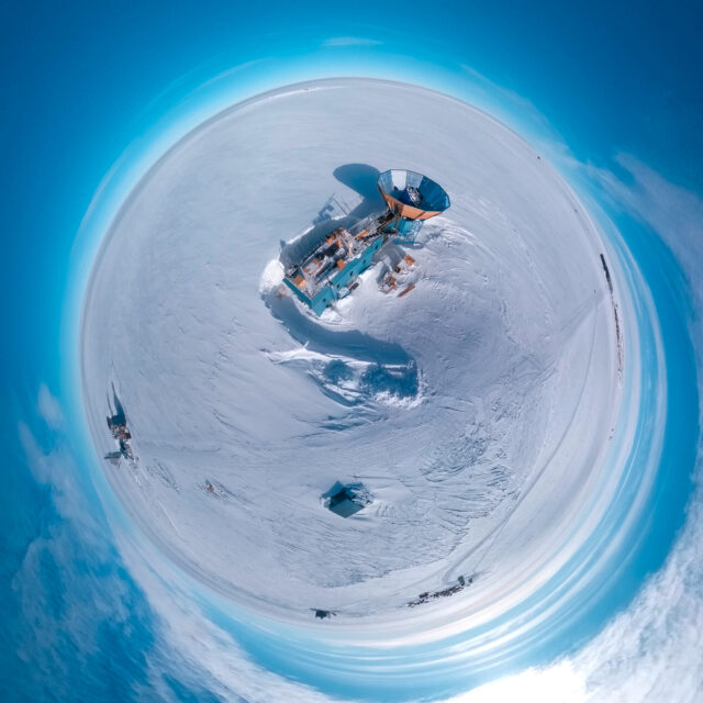 Aerial "little planet" view of a two-story blue building on an ice sheet, with a tower with a circular metal cone-shaped ground shield on top and a radio telescope array inside it.