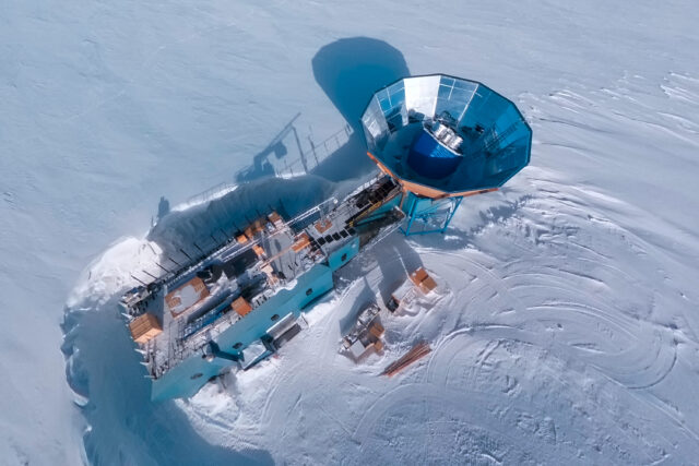 Aerial view of a two-story blue building on an ice sheet, with a tower with a circular metal cone-shaped ground shield on top and a radio telescope array inside it. The building is in a depression, with the snow level significantly higher than the base of the building.
