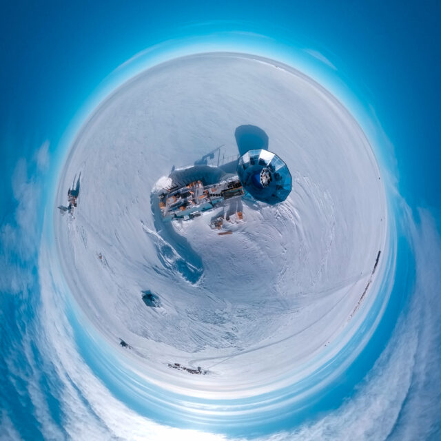 Aerial "little planet" view of a two-story blue building on an ice sheet, with a tower with a circular metal cone-shaped ground shield on top and a radio telescope array inside it.