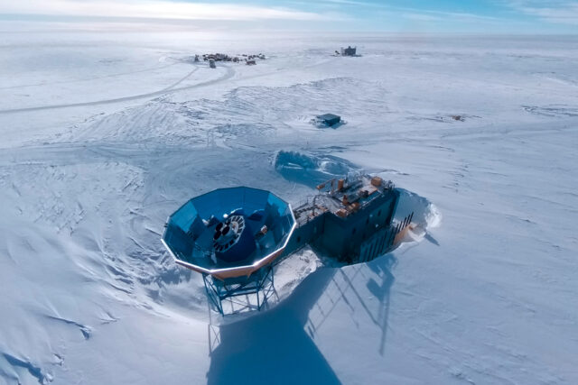 Aerial view of a two-story blue building on an ice sheet, with a tower with a circular metal cone-shaped ground shield on top and a radio telescope array inside it. The building is in a depression, with the snow level significantly higher than the base of the building.