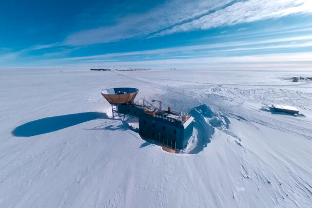 Aerial view of a two-story blue building on an ice sheet, with a tower with a circular metal cone-shaped ground shield on top and a radio telescope array inside it. The building is in a depression, with the snow level significantly higher than the base of the building.