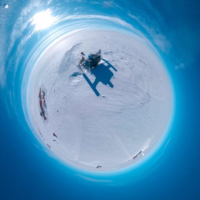 Aerial "little planet" view of a two-story blue building on an ice sheet, with a round metal-clad tower connected to each end of the building with a bridge.